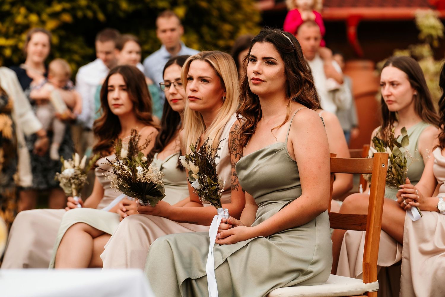 Bridesmaids in neutral-colored dresses seated during outdoor wedding ceremony.