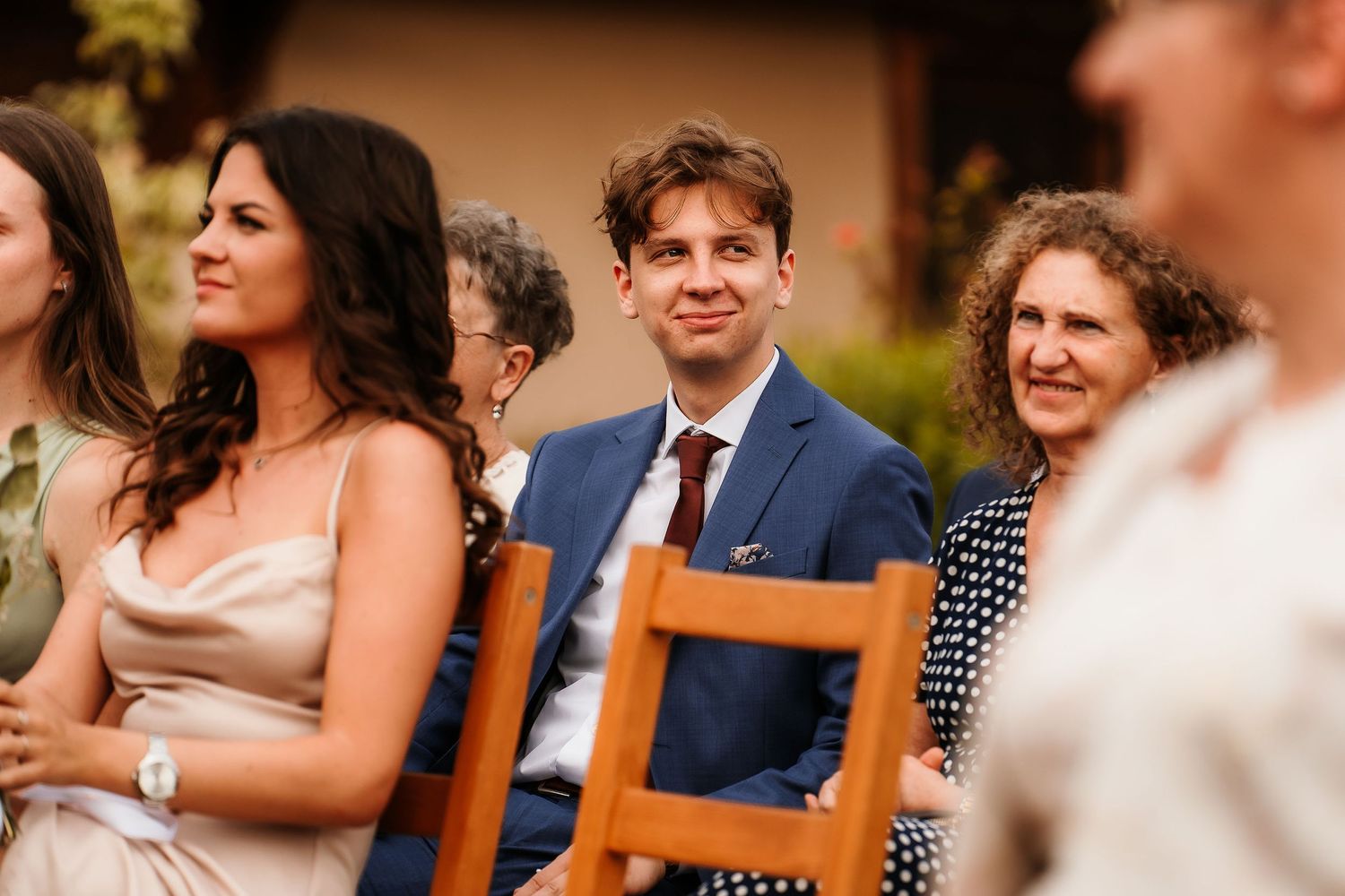 Wedding guests seated on wooden chairs during outdoor ceremony.