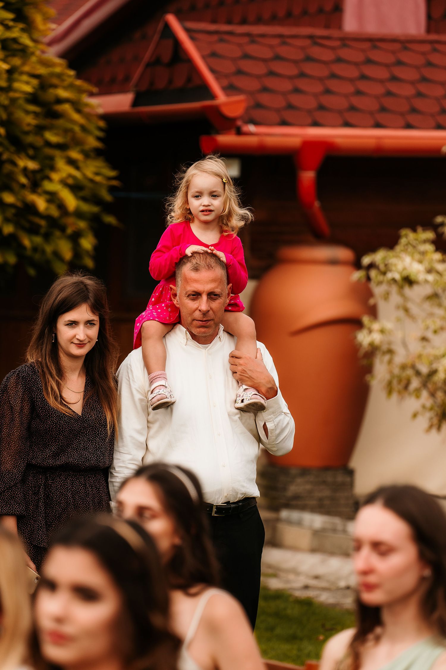 Young child in bright red dress sits on shoulders during outdoor wedding celebration.