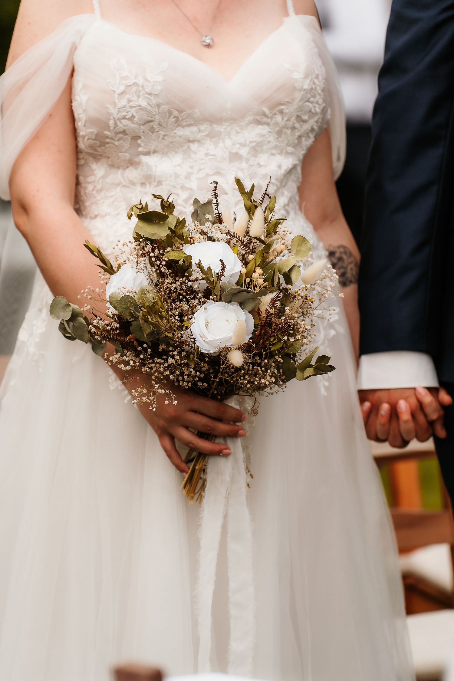 Close-up of bridal bouquet with white roses and dried flowers against white wedding dress.
