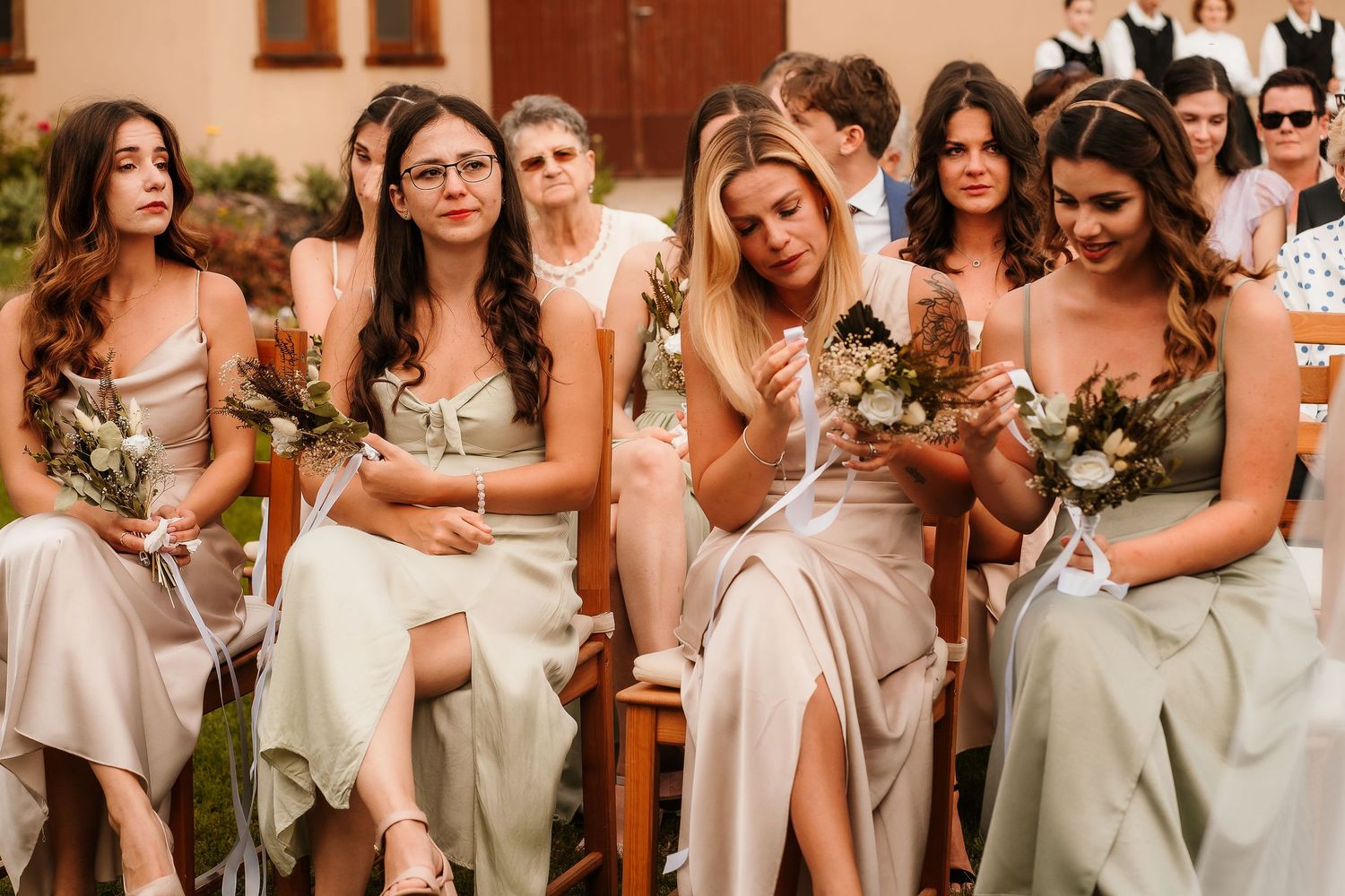 Bridesmaids in sage green dresses sitting together during outdoor wedding ceremony.