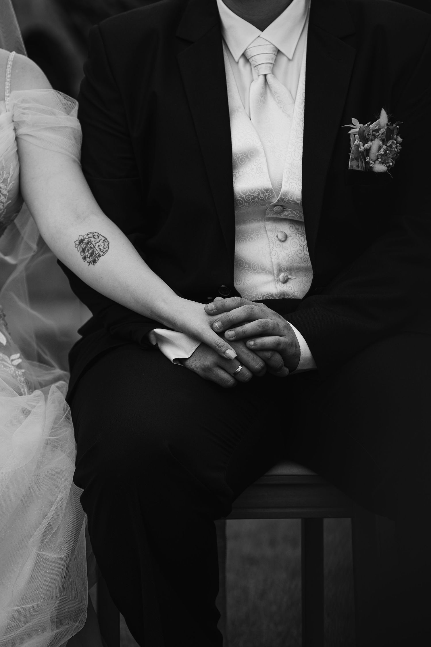 Black and white detail shot of couple holding hands, showing wedding attire and tattoo.