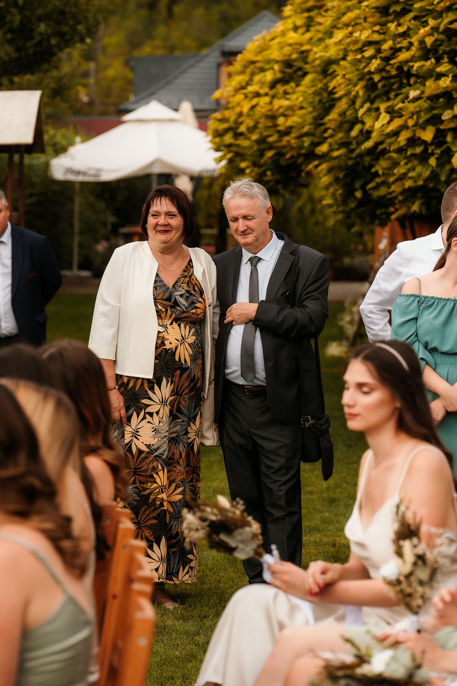 Wedding guests in formal attire gather on a lawn with autumn foliage in the background.