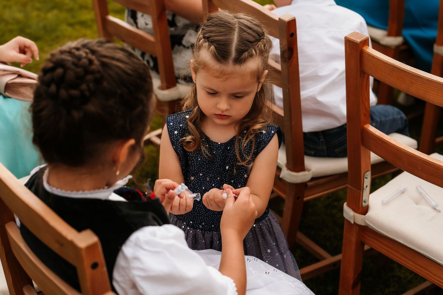 Two young children in formal wear interact while seated in wooden chairs at an outdoor event.