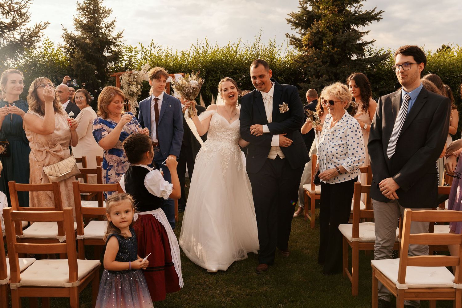 Wedding party and guests gather on a lawn with wooden chairs during an outdoor ceremony.