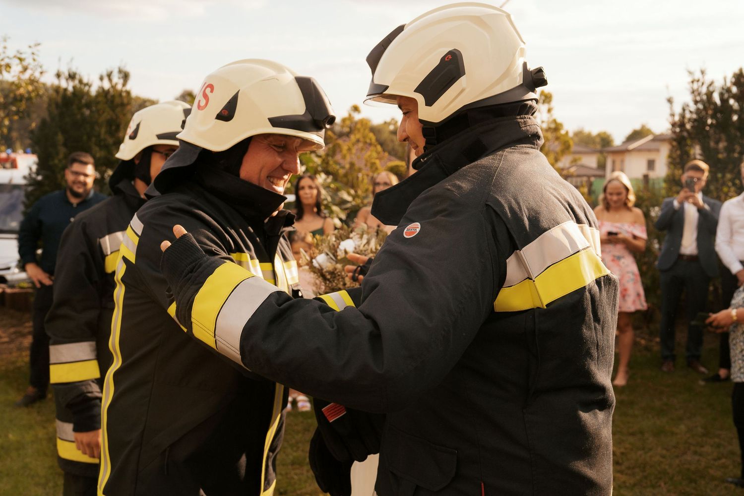 Two firefighters in protective gear and helmets interact at an outdoor event.