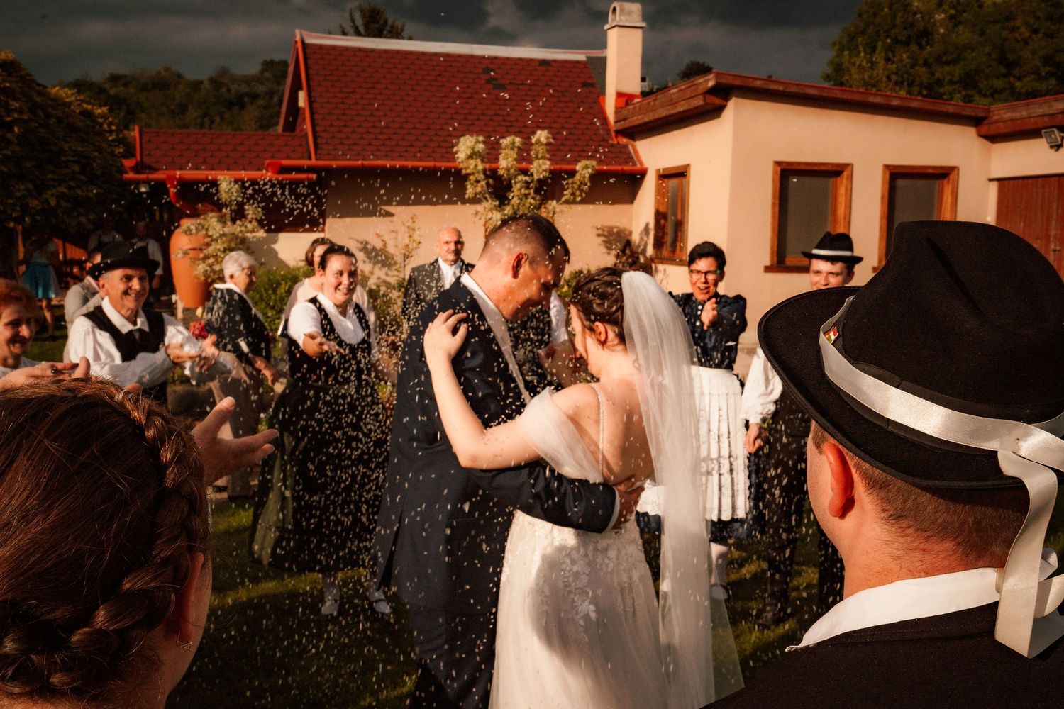 Wedding celebration with water spray against sunset backdrop near red-roofed building.