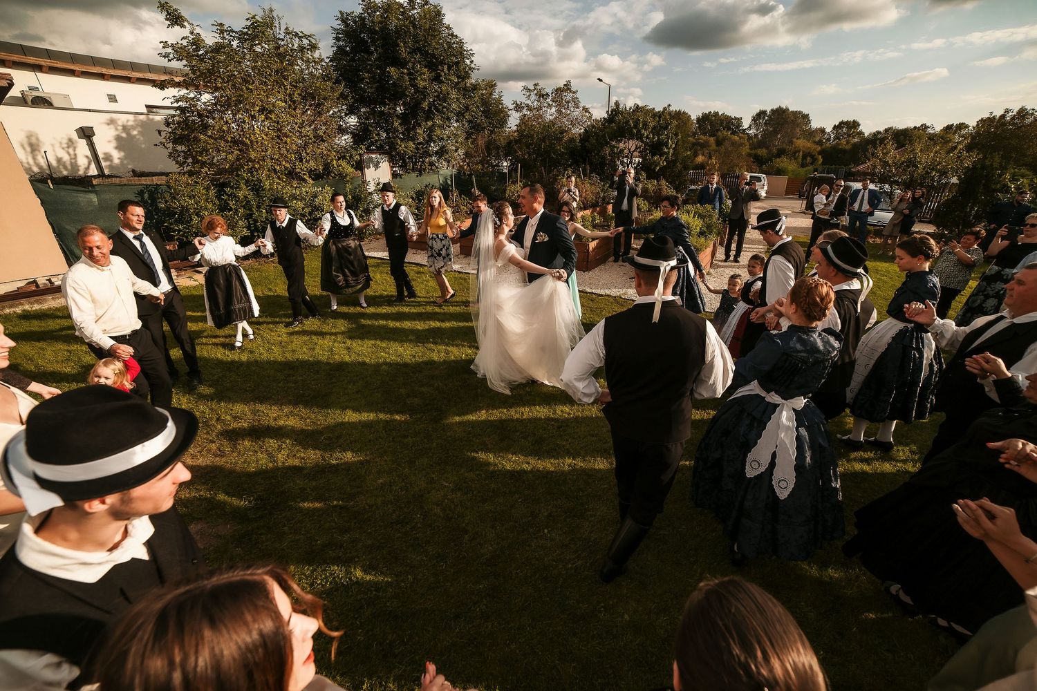 Wedding celebration with guests dancing in circular formation on outdoor lawn.