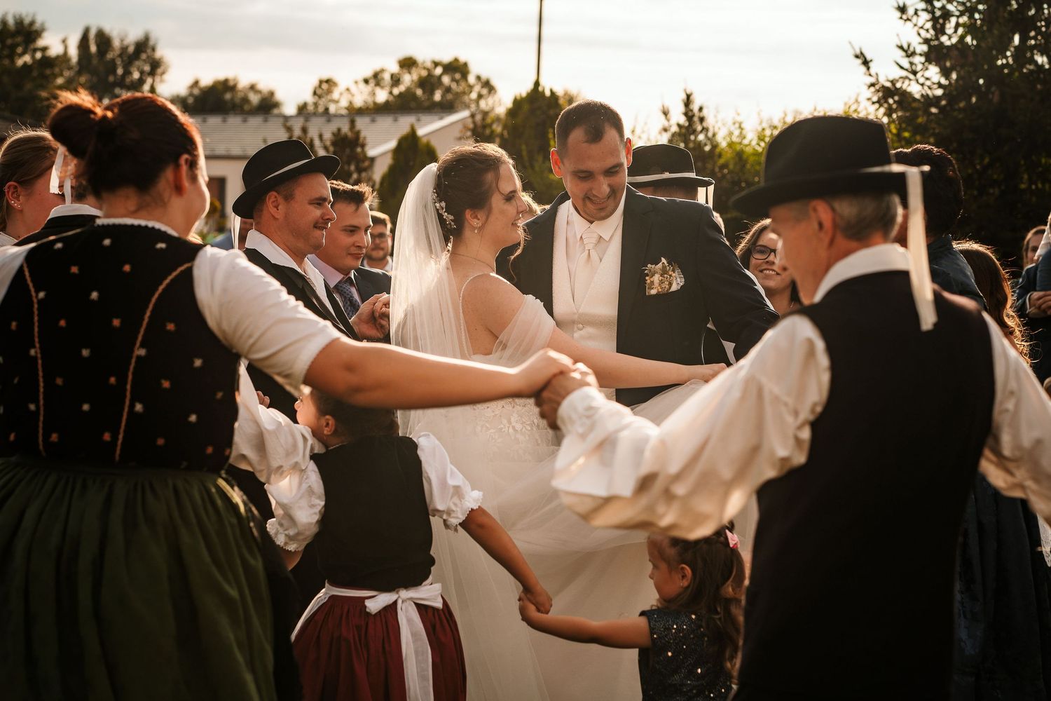 Traditional folk dancers in period costumes celebrate at an outdoor wedding reception during sunset.