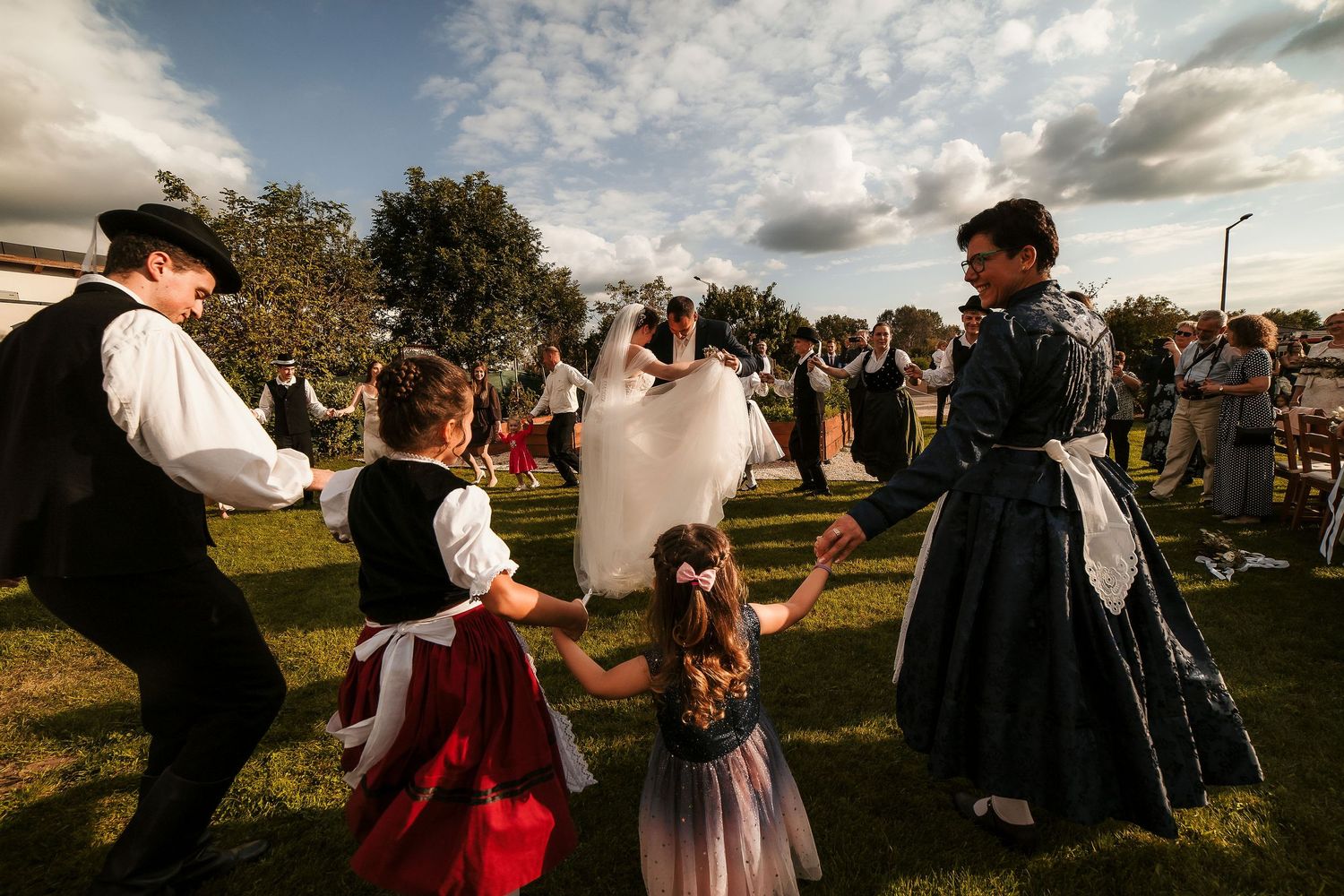 Folk dancers in traditional dress perform at an outdoor wedding celebration under a dramatic sky.