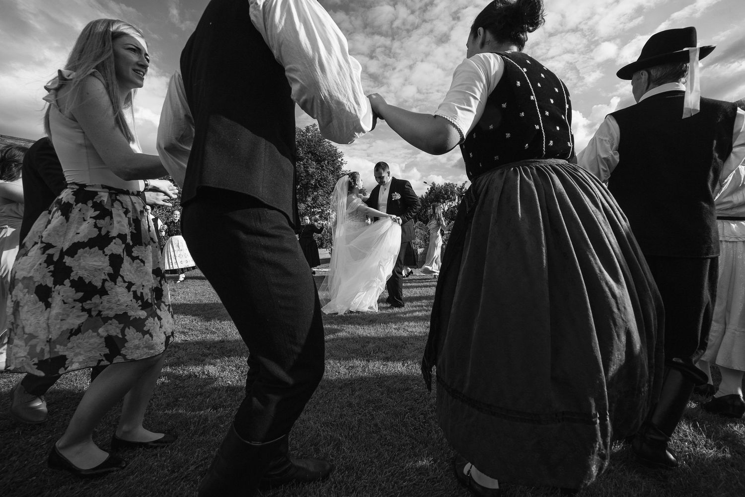 Black and white photo of traditional folk dancing at a wedding celebration outdoors.