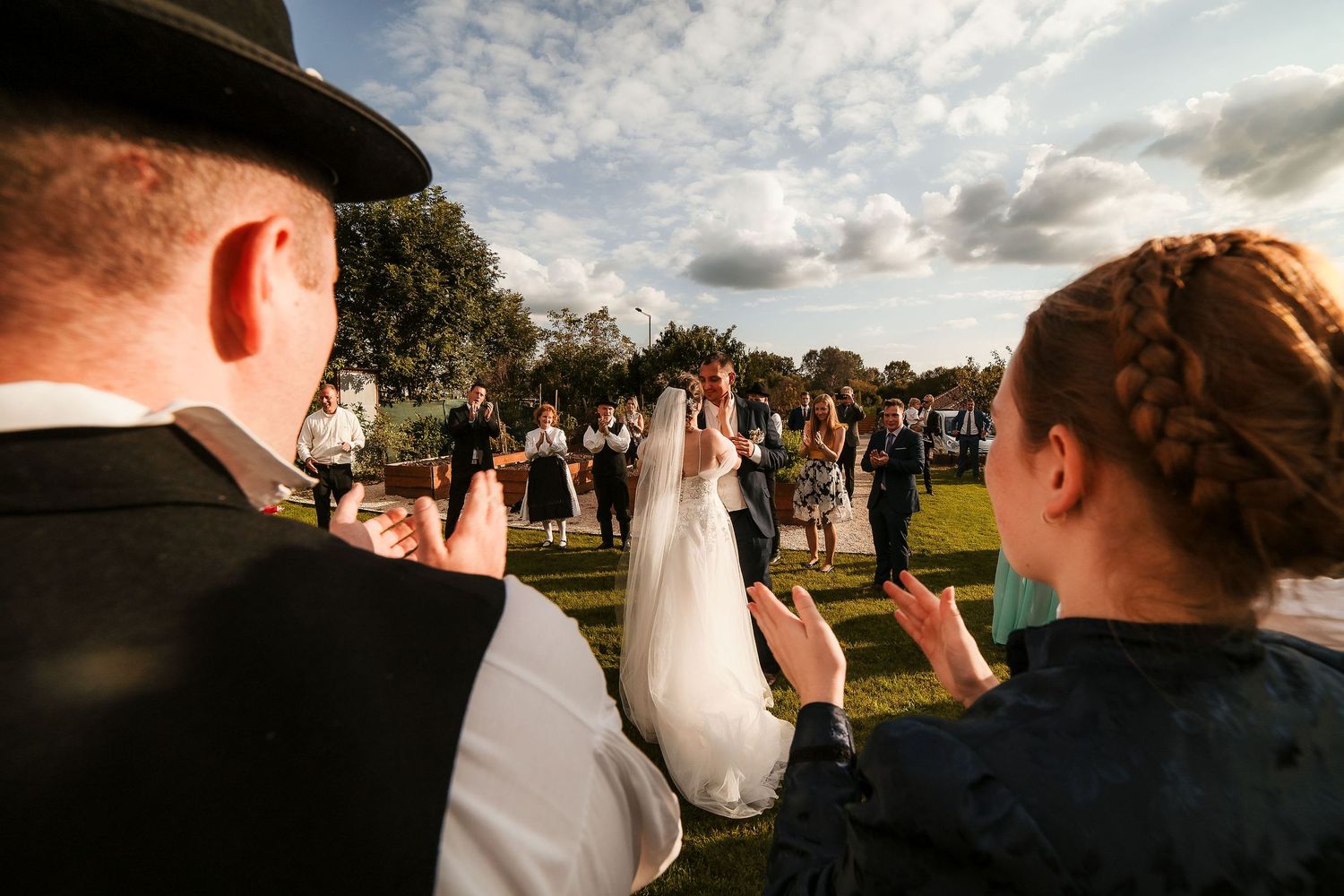 Guests in traditional dress photograph a wedding celebration outdoors.