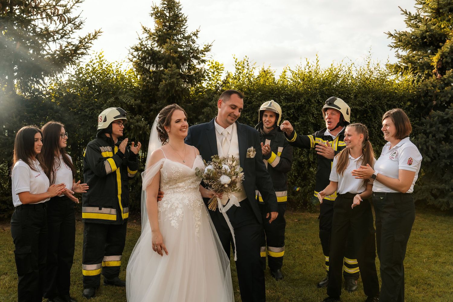 Wedding party poses with firefighters in uniform during an outdoor celebration.