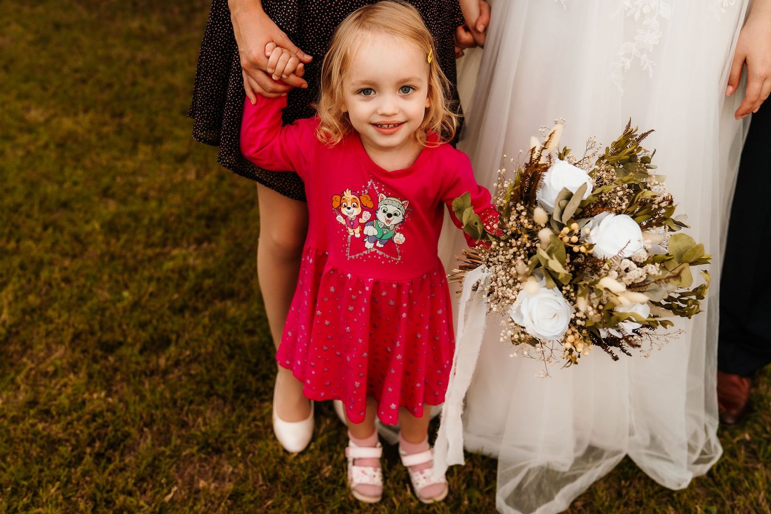 A young girl in a bright red dress stands next to a wedding bouquet featuring white flowers and greenery.