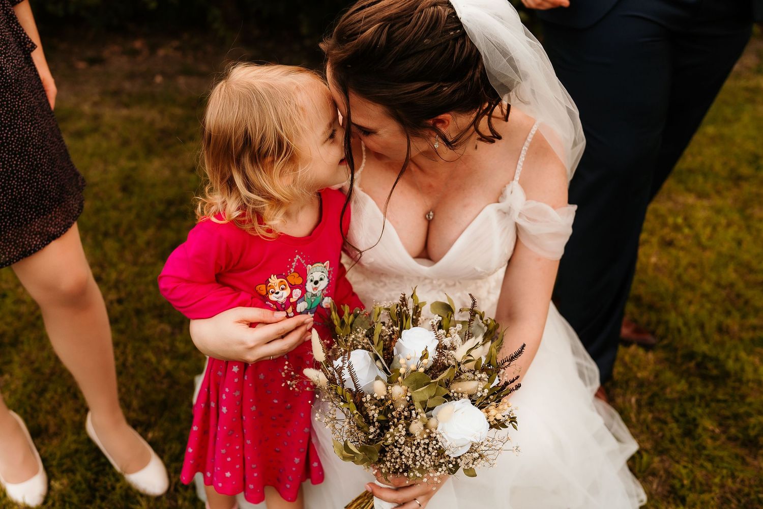 A bride in white dress holds a rustic bouquet while sharing a tender moment with child in red dress.