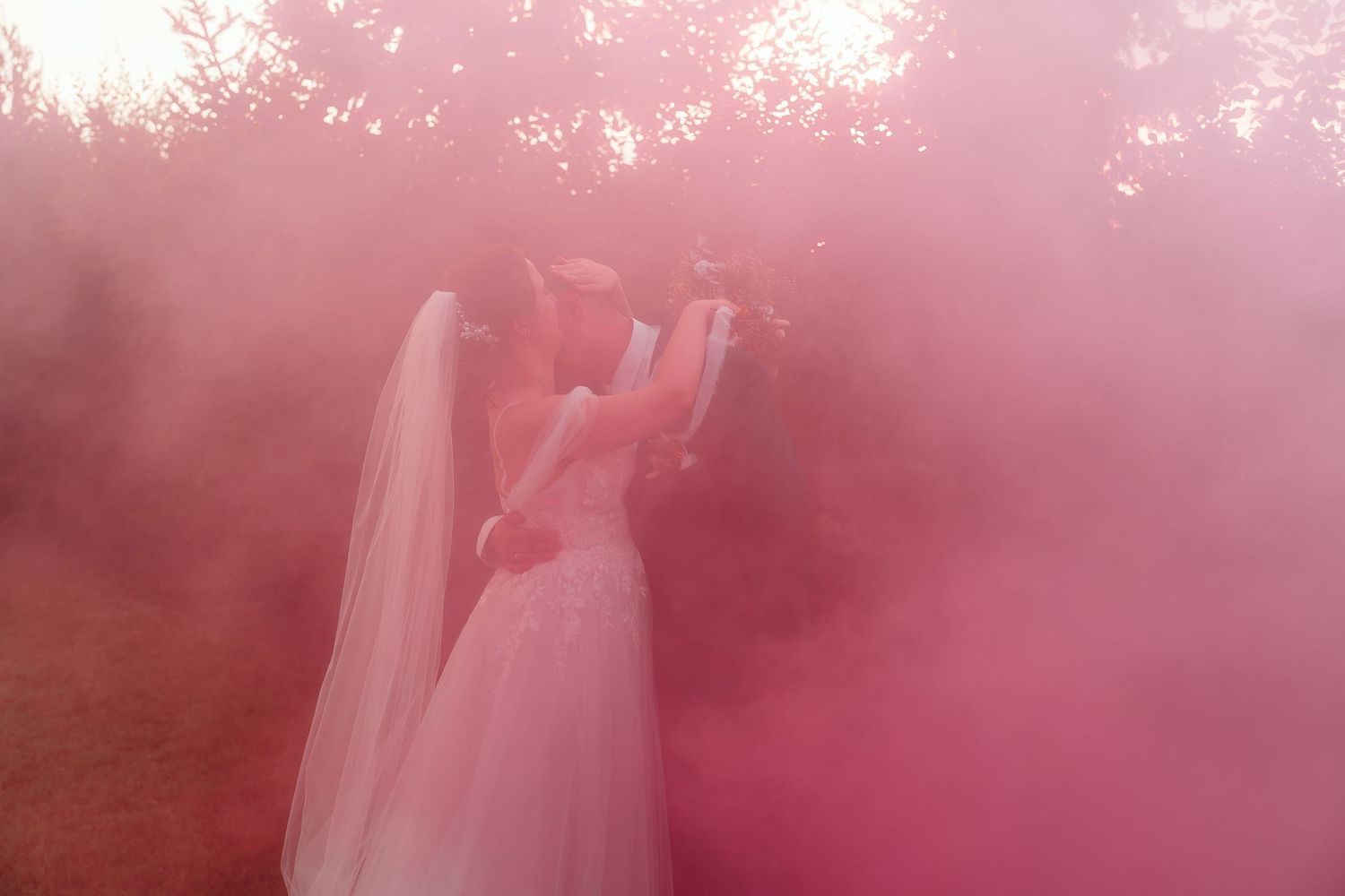Newlyweds share an intimate moment surrounded by pink smoke in an outdoor setting.