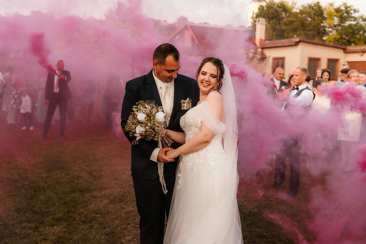 A couple poses together amid vibrant pink smoke during their outdoor wedding celebration.