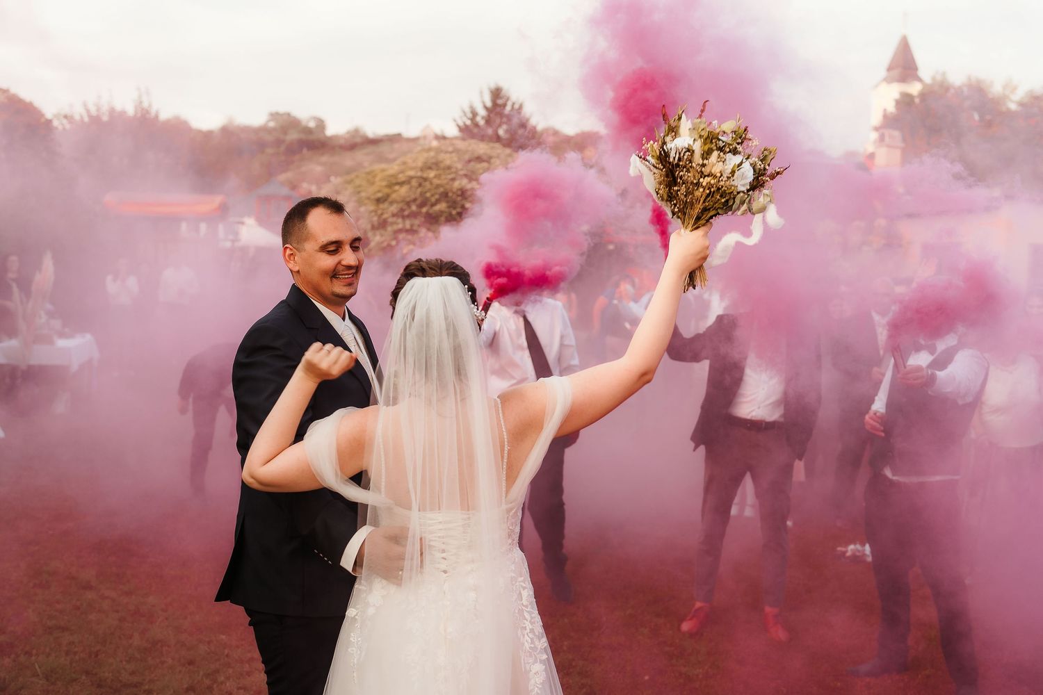 Newlyweds celebrate amid pink smoke clouds during their outdoor wedding reception.