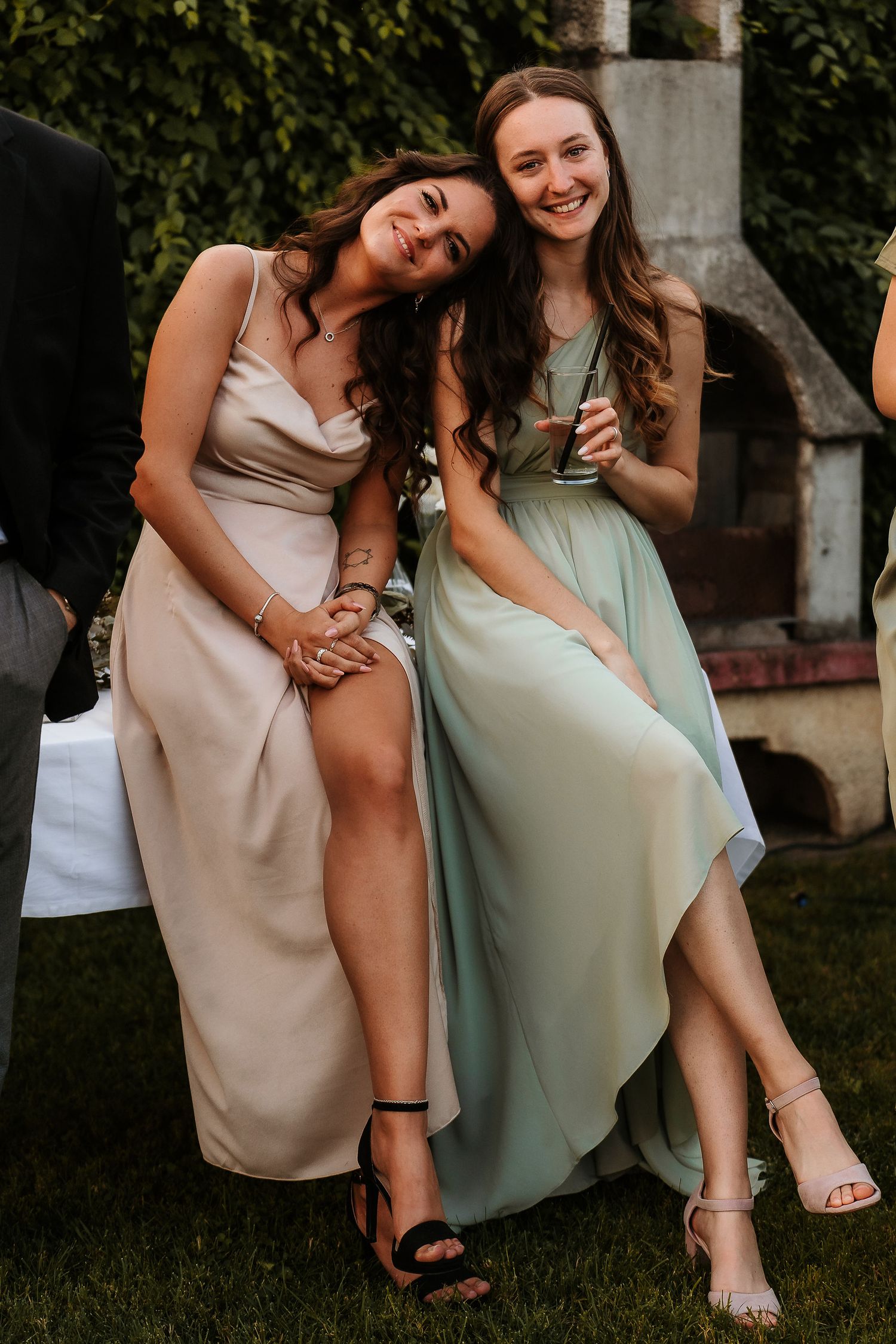 Two wedding guests in elegant dresses share a candid laughing moment on the lawn.