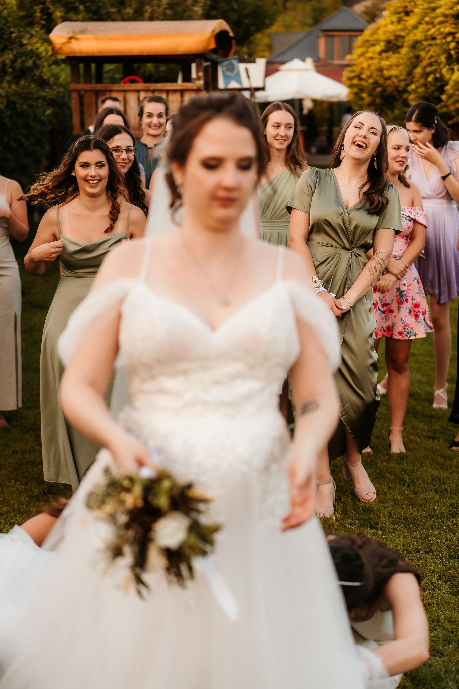 Wedding guests in sage green dresses laugh joyfully behind a bride in a white gown at an outdoor garden ceremony.