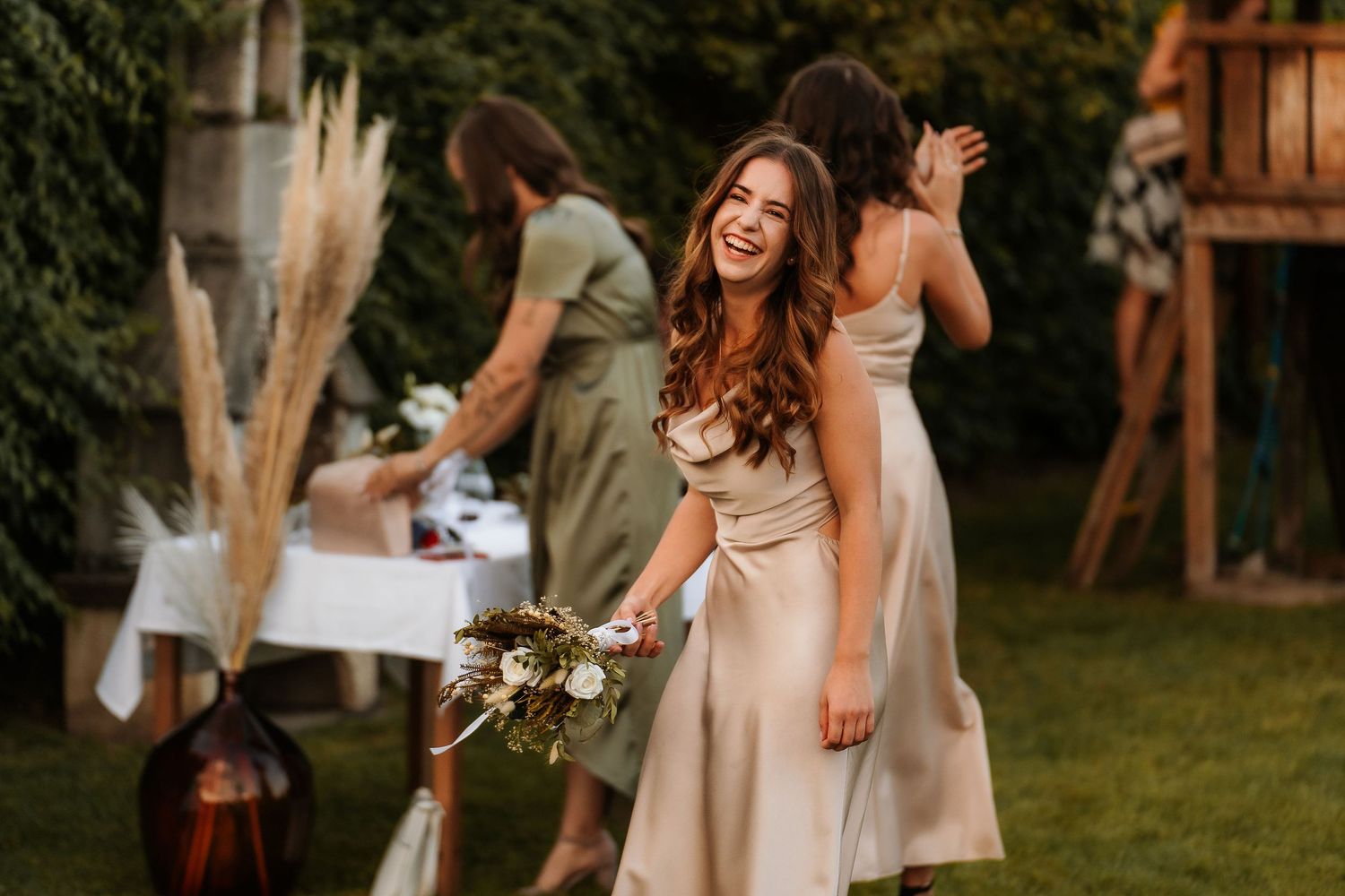 Bridesmaid in champagne dress holds bouquet while laughing during outdoor celebration.