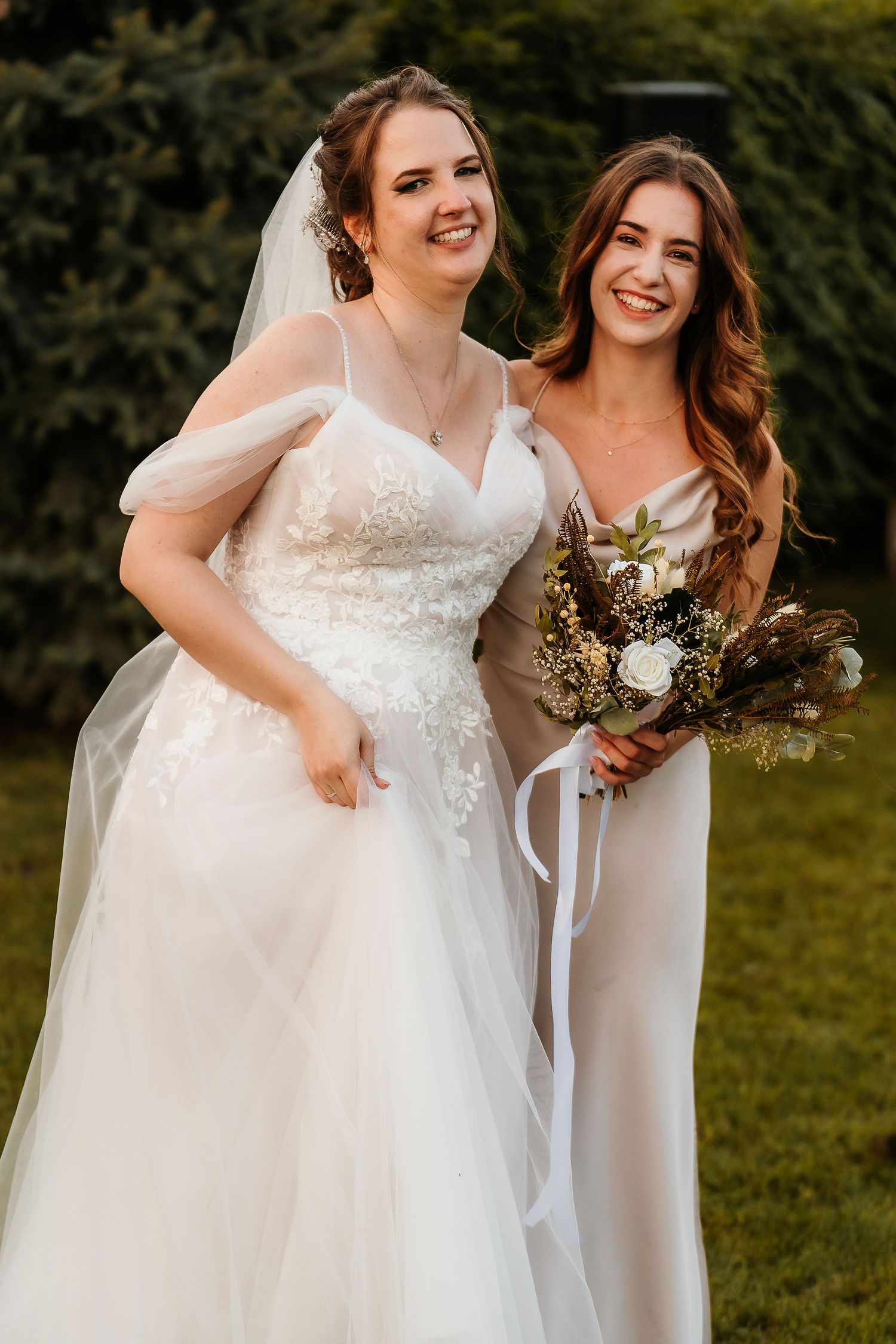 Happy bride in lace gown poses with bridesmaid holding rustic bouquet at outdoor wedding.