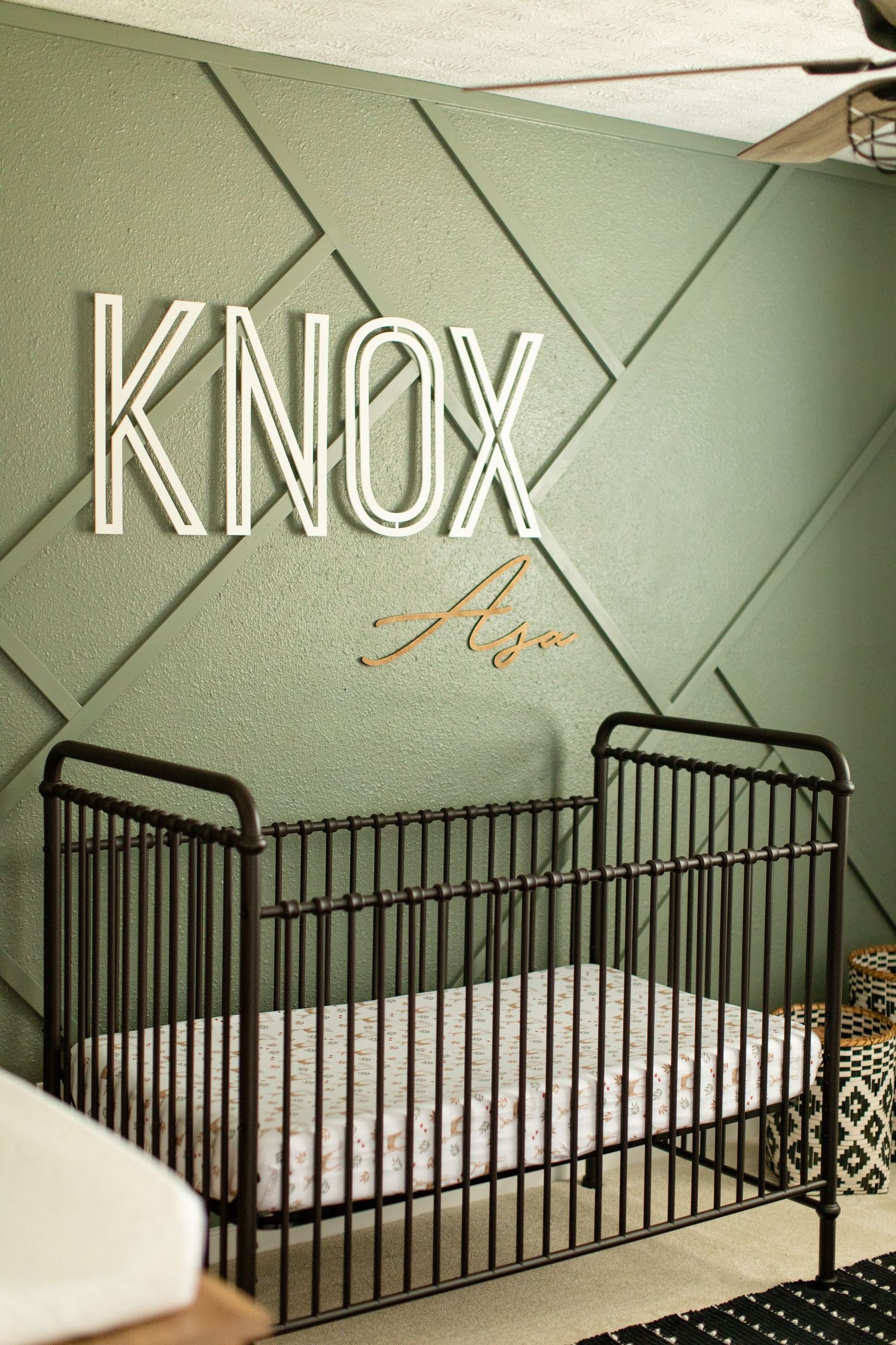 A baby nursery wall with the name Knox displayed in modern lettering above a black metal crib.