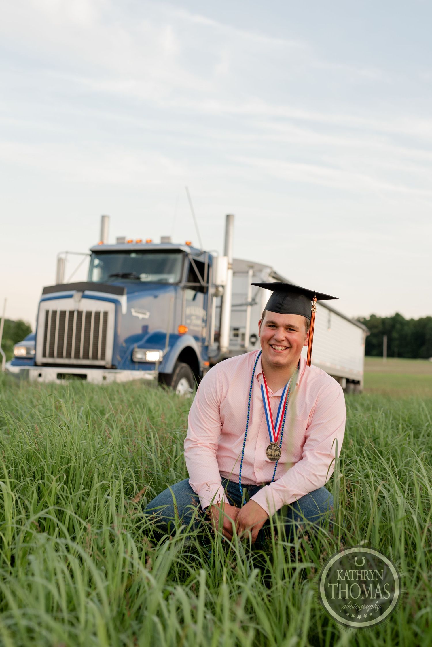 Graduate sitting in tall grass with semi truck in background wearing pink shirt and graduation cap.