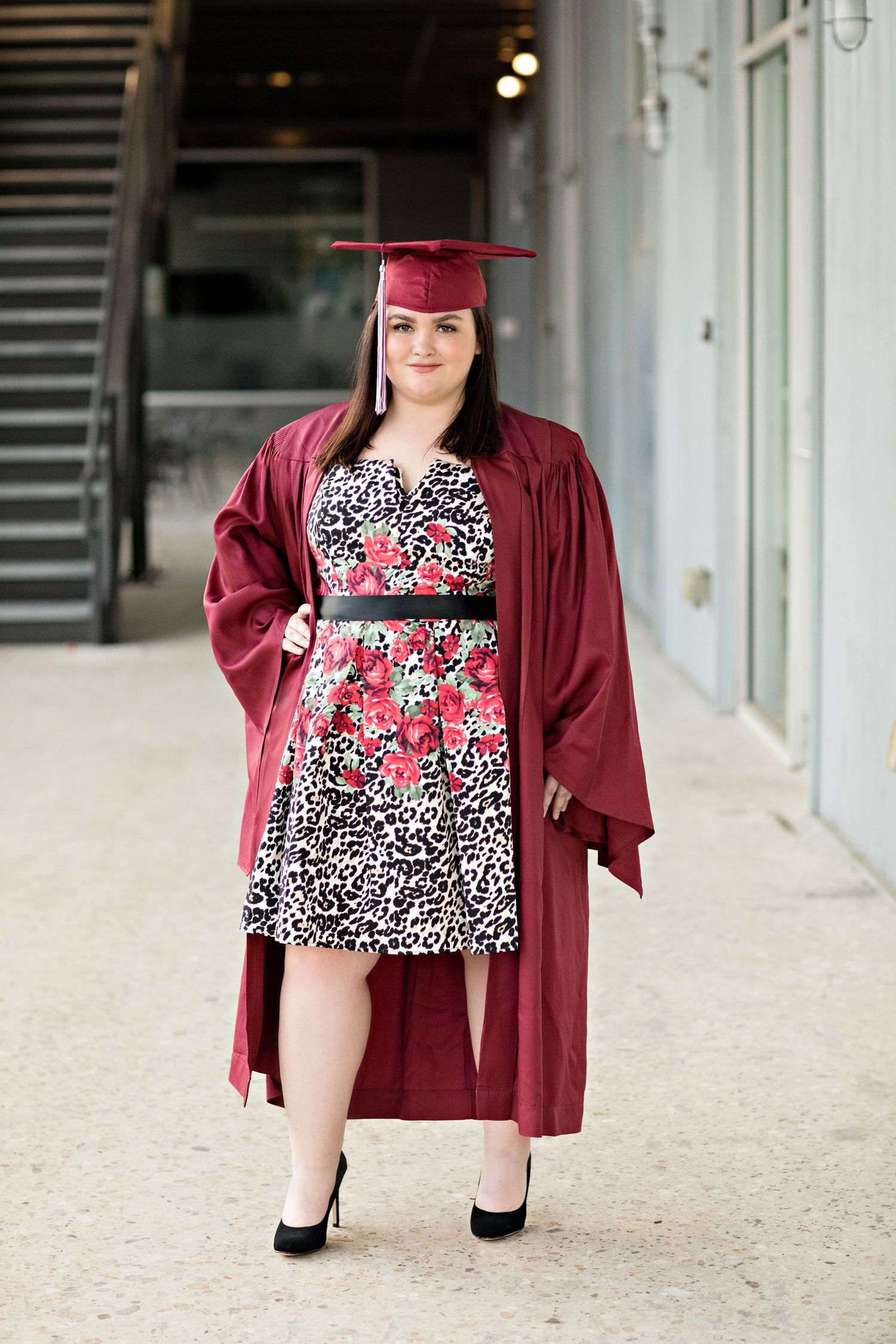 Graduate in maroon cap and gown wearing a floral print dress poses for graduation photos.