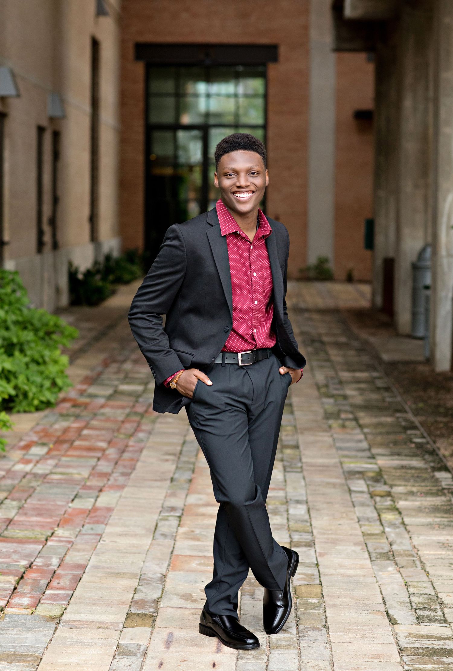Professional portrait of person in dark suit and red shirt posing in brick courtyard.