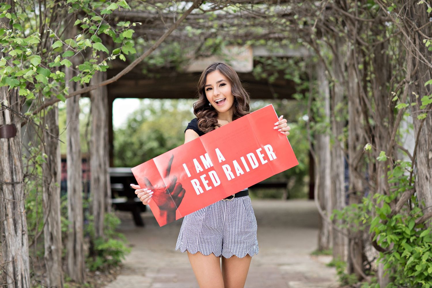 Student holding red graduation banner in garden archway.