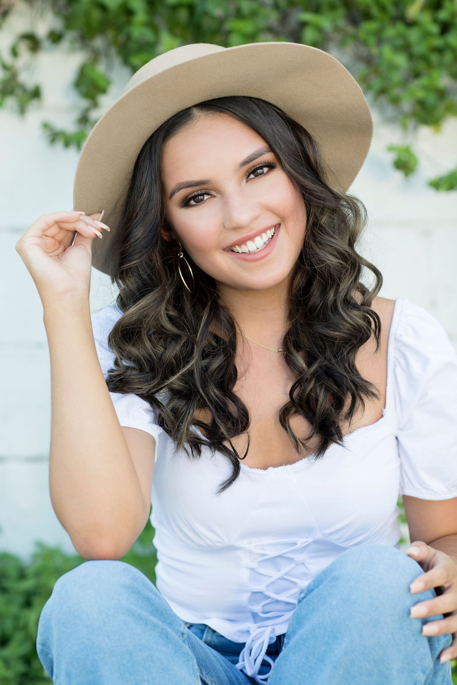 Casual portrait in white top and denim with wavy hair and tan hat against greenery.