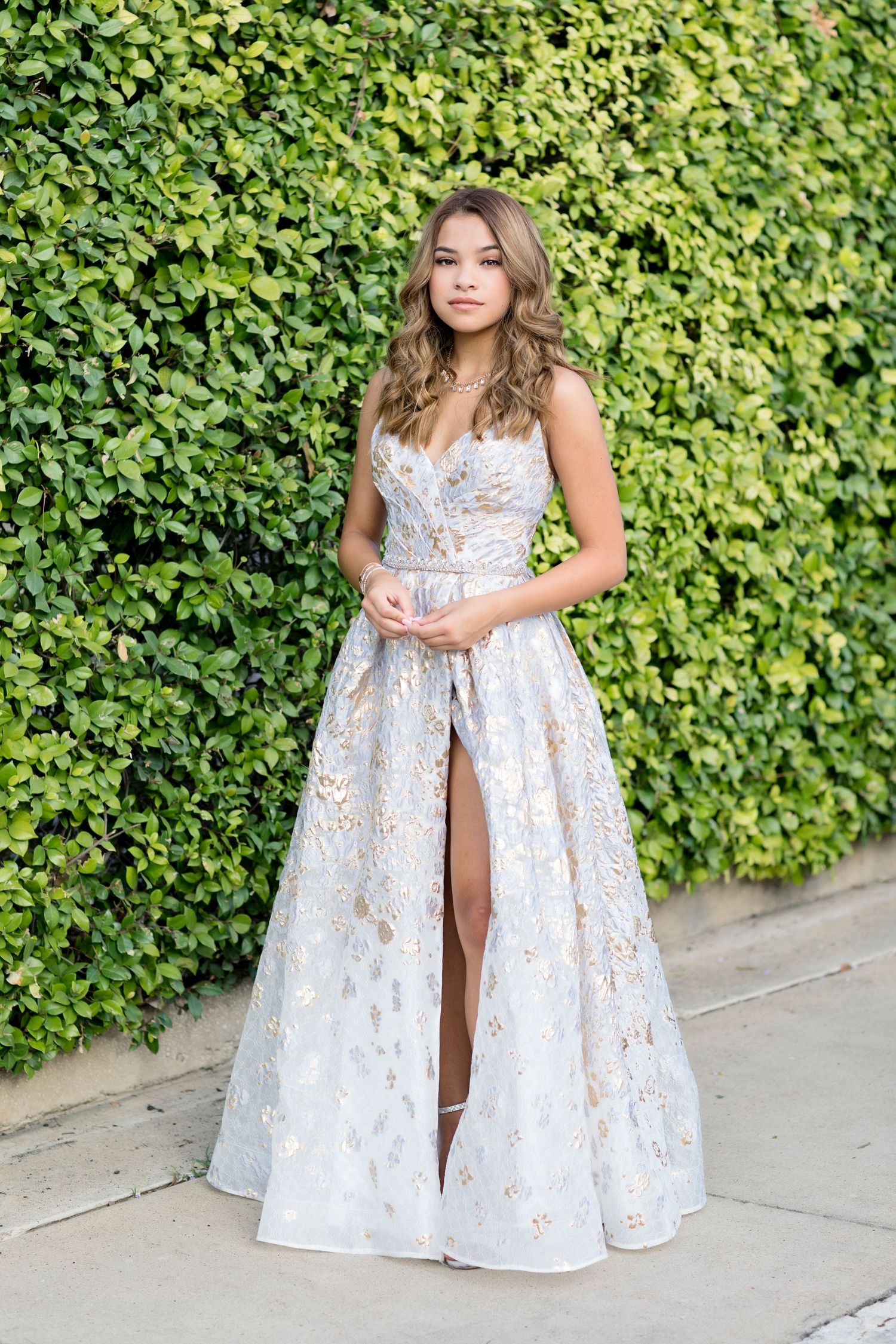 Elegant portrait in white and silver formal gown with slit against green hedge wall.