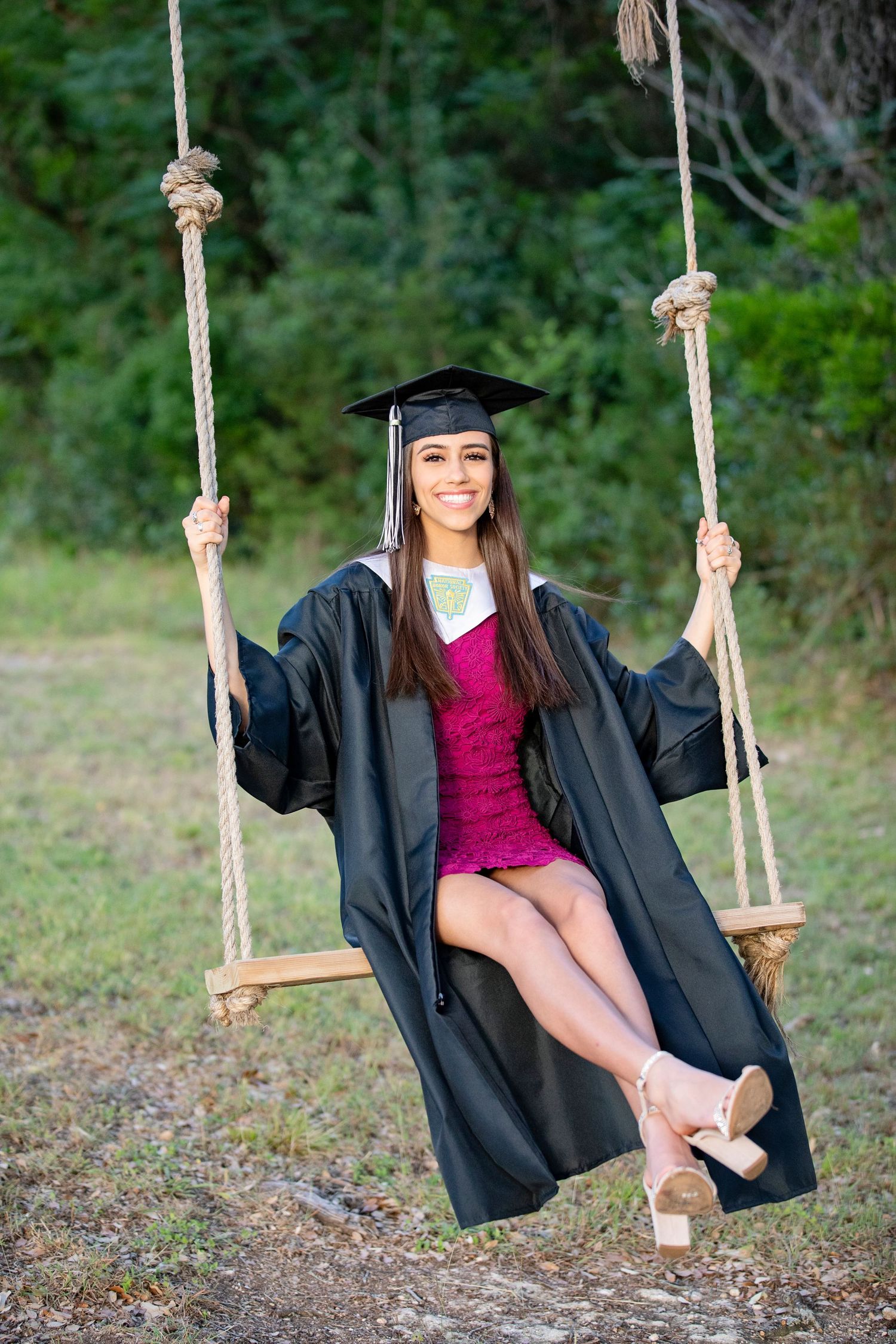 Graduate in black cap and gown sitting on rustic rope swing outdoors.