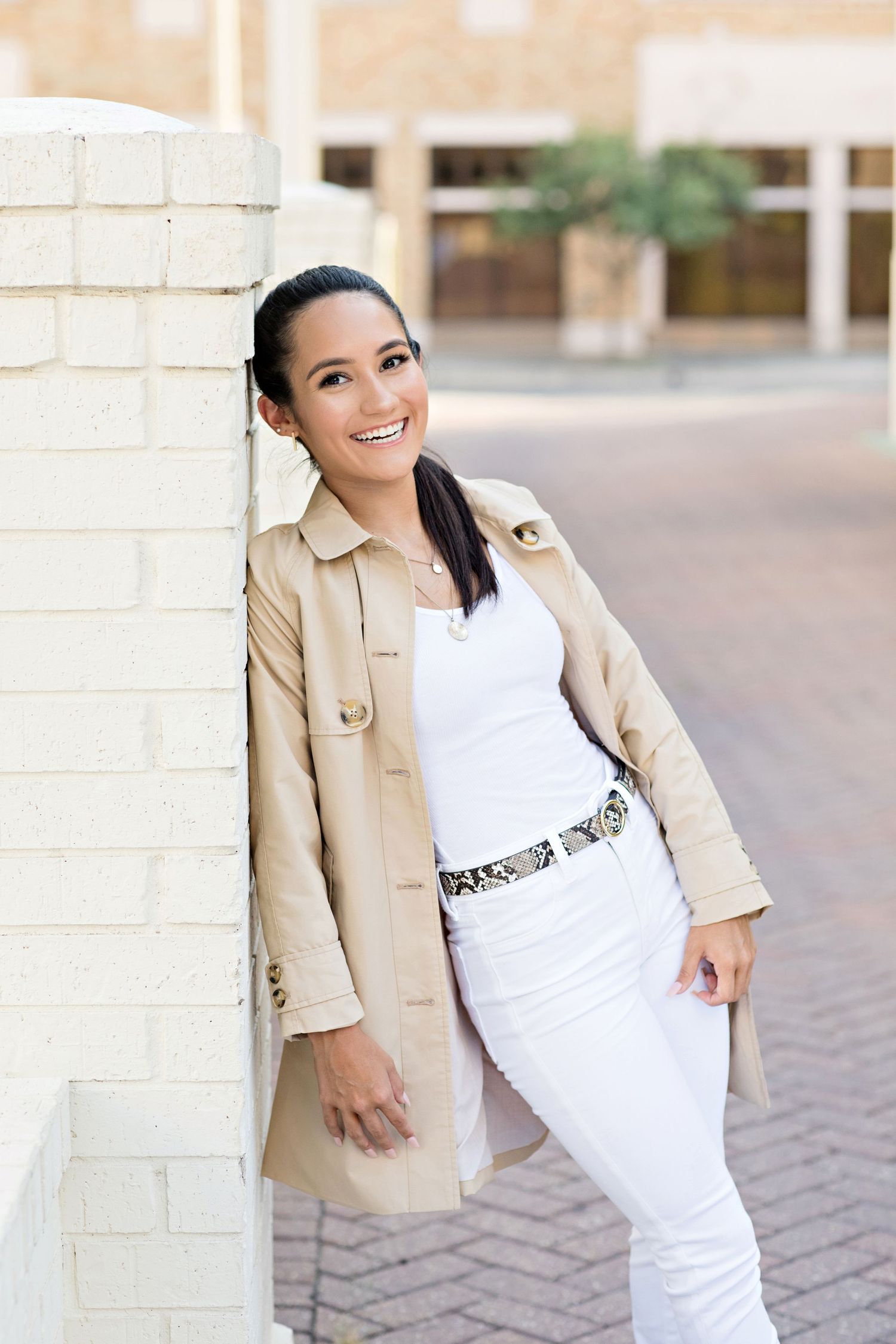 Professional portrait of someone in a beige trench coat and white outfit against a brick wall.