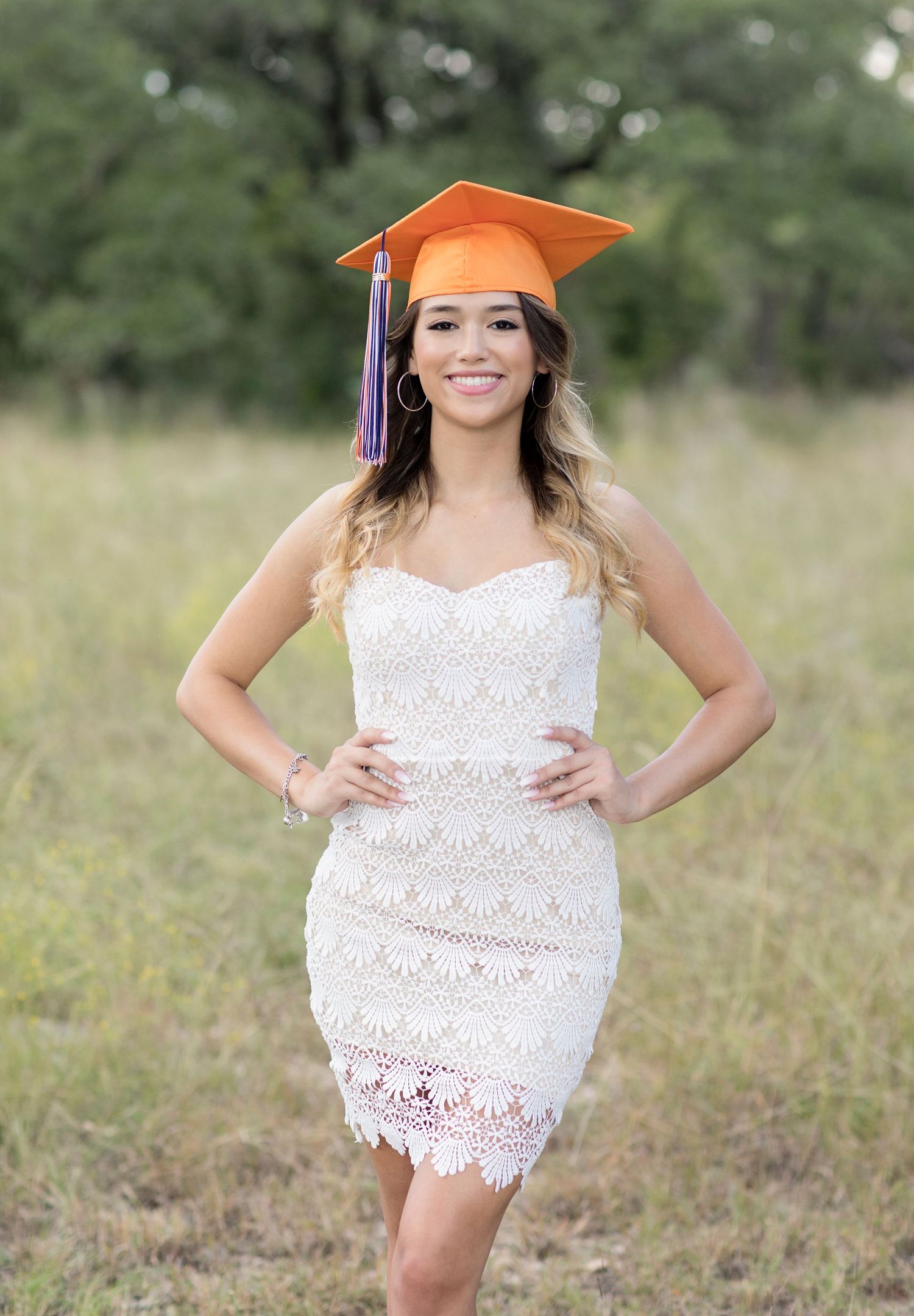 Graduate in white lace dress and orange cap posing in field.
