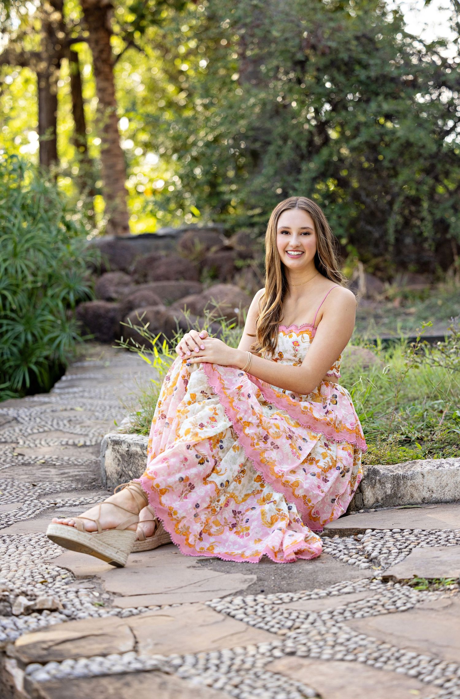A person in a floral pink dress sits on a decorative stone pathway in a garden setting with natural sunlight.