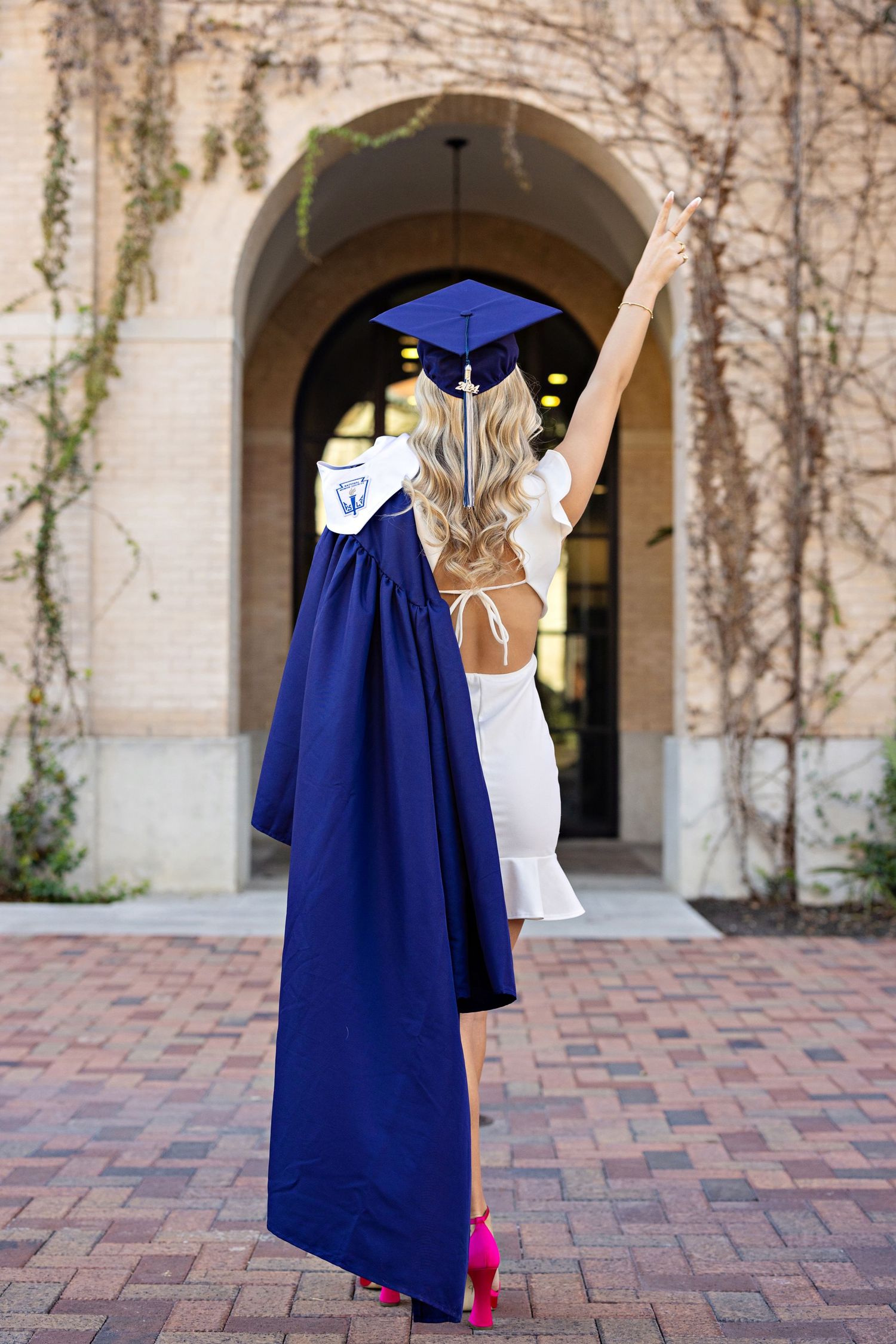 Graduate in blue cap and gown celebrates with raised arms while wearing a white dress and bright pink heels.