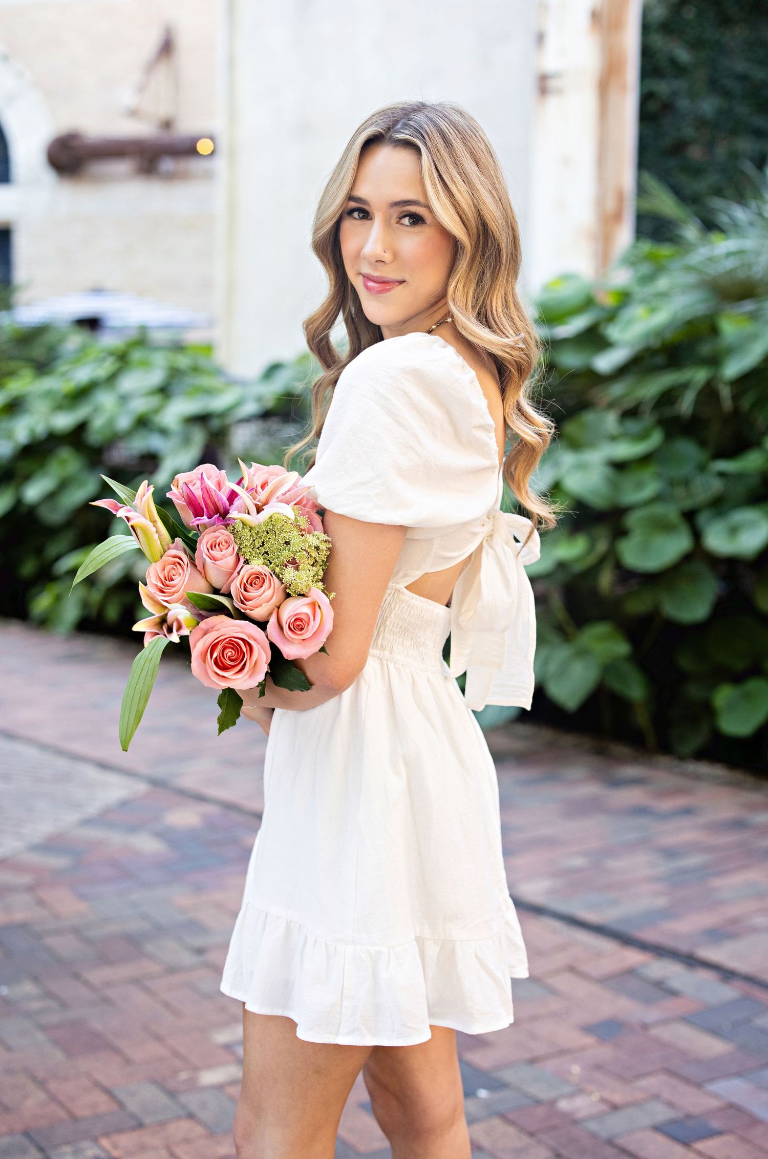 A woman in a white summer dress with bow detail holds a bouquet of pink and coral roses on a brick pathway.
