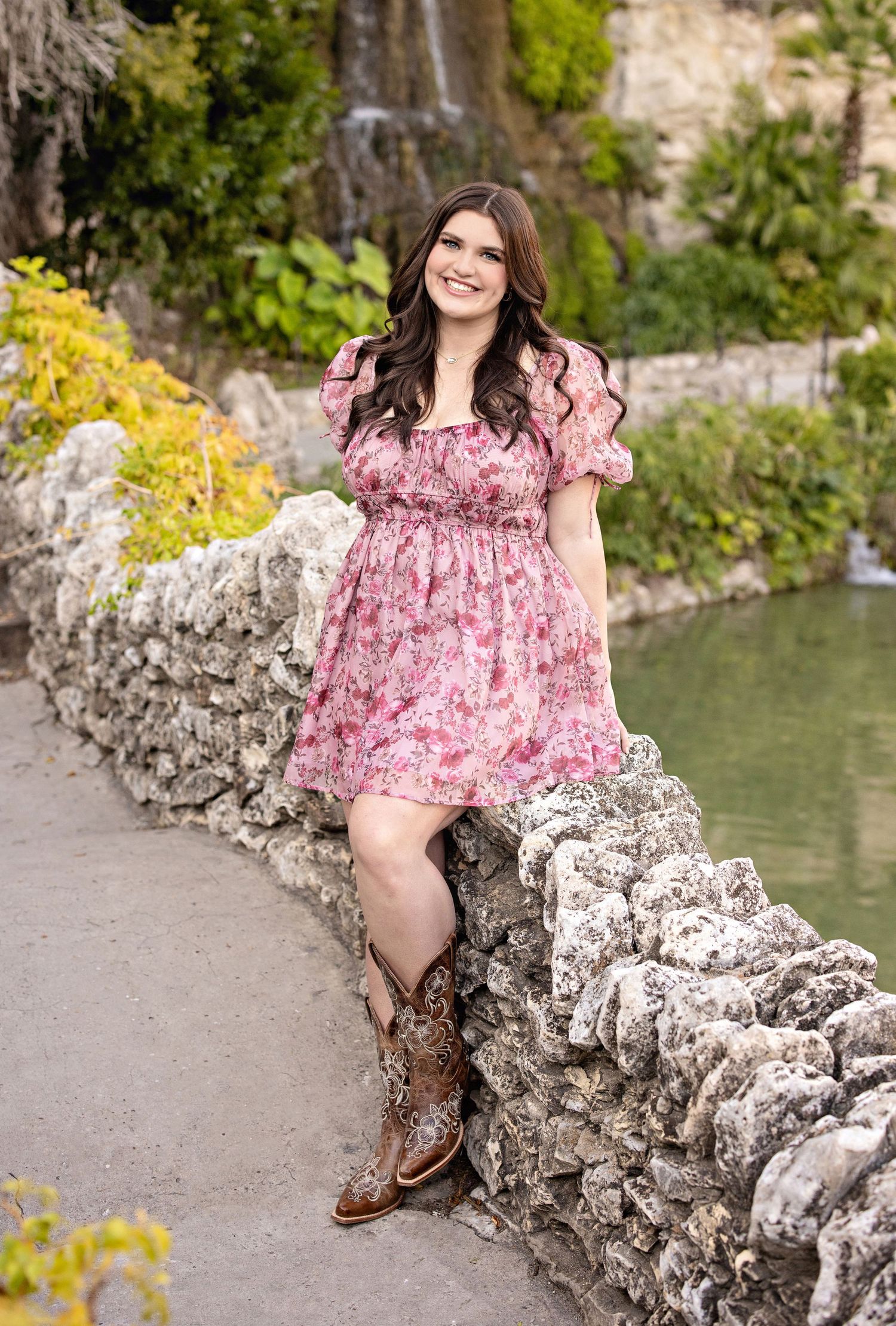 A pink floral dress with cowboy boots creates a country chic look against a stone wall and waterfall backdrop.