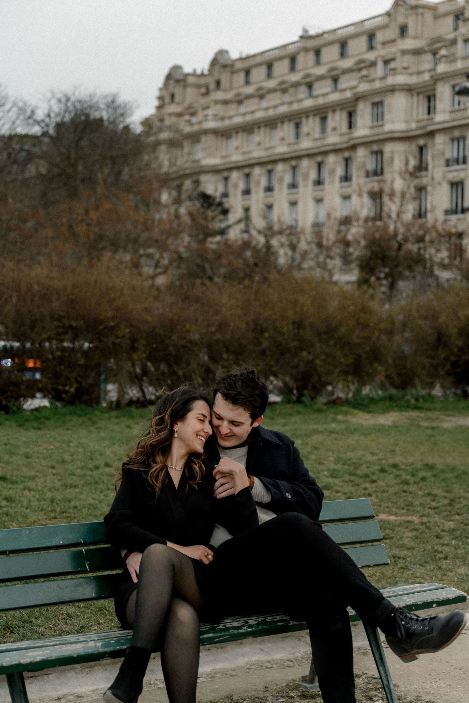 A romantic couple sits close together on a park bench in front of a grand Parisian building.