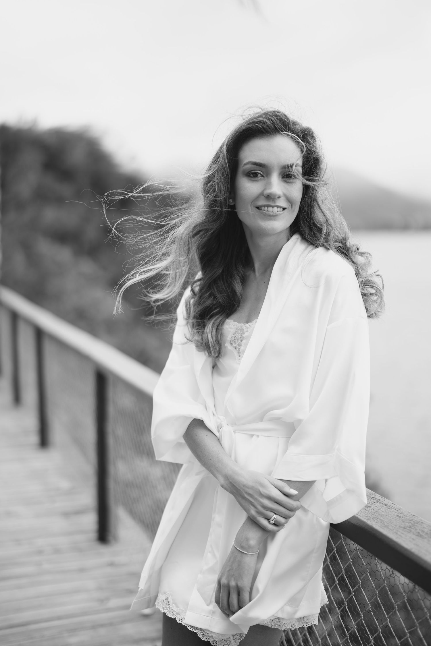 Woman in flowing white dress stands on coastal boardwalk during sunset.