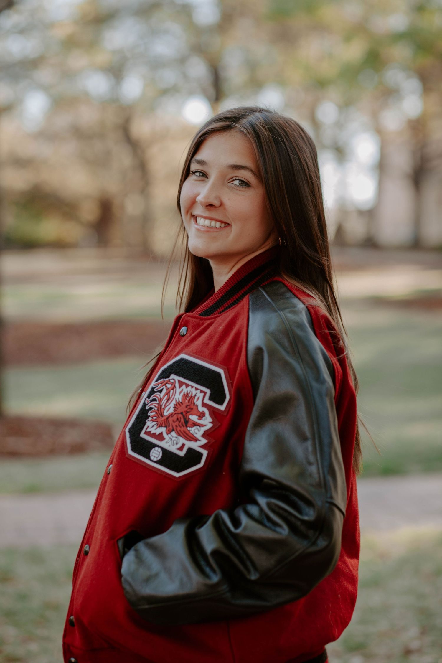 College graduate and volleyball student-athlete poses in a nature landscape with her letterman jacket.