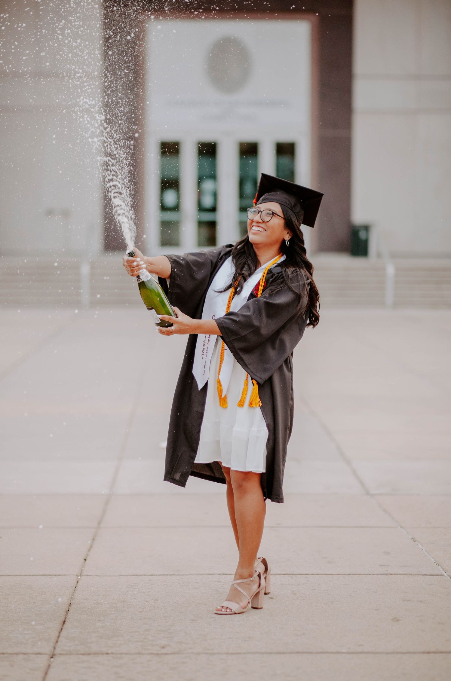 First generation college graduate smiles big as she pops a bottle of champagne in front of her college.