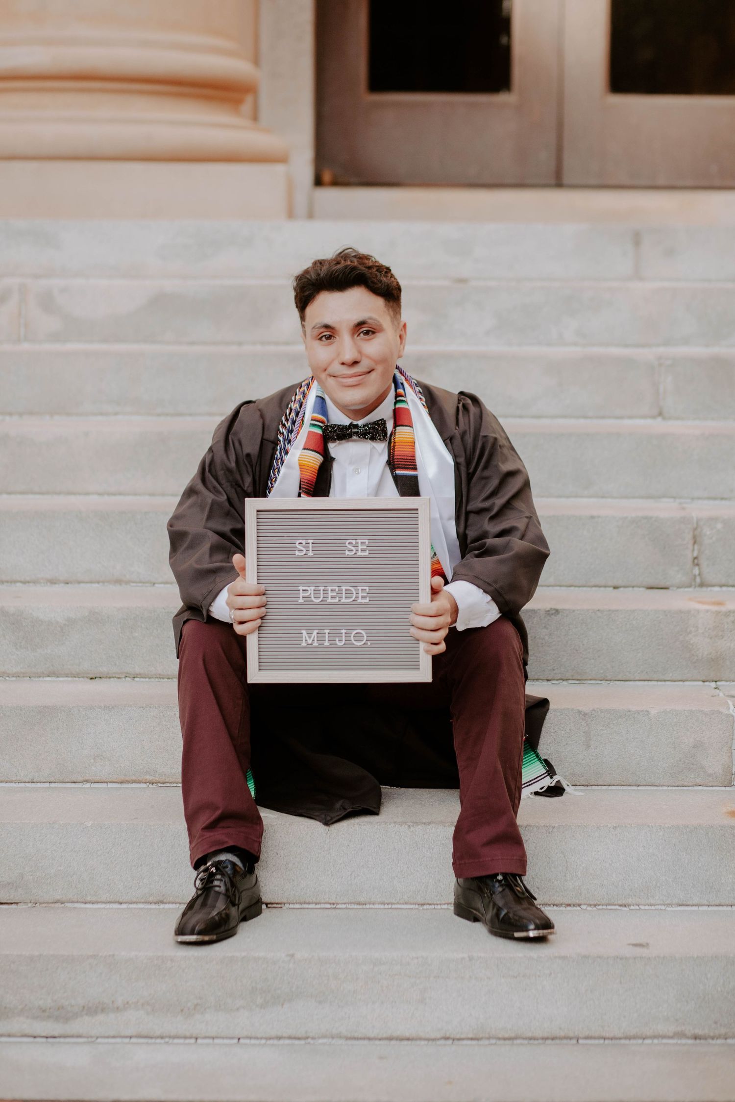 College student poses on the stairs with a sign that says "Si se puede, mijo".