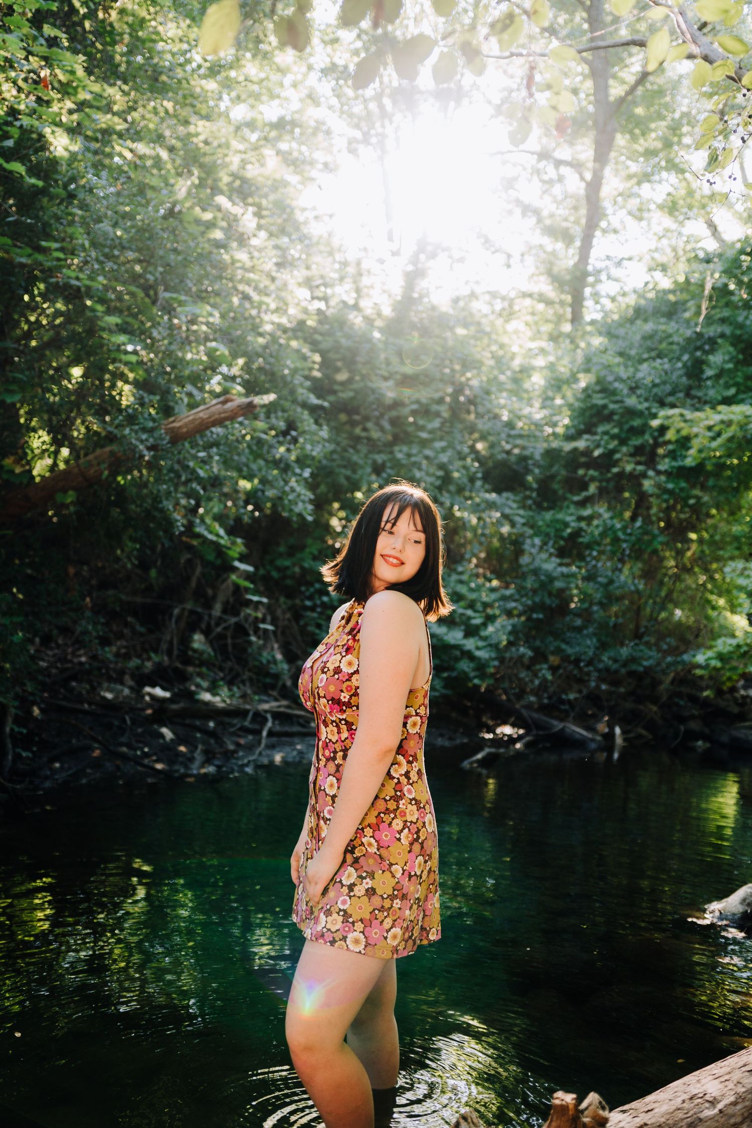 Someone in a floral top poses playfully at the edge of a serene lake surrounded by lush greenery.