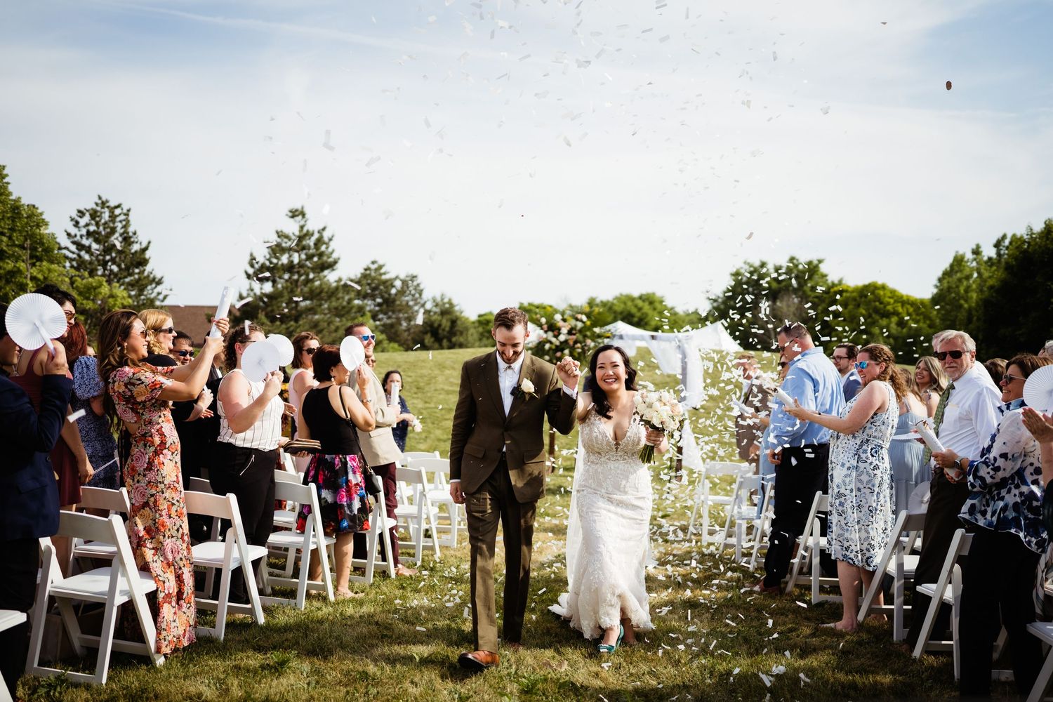Wedding guests throw confetti as the newlyweds walk down the aisle at an outdoor ceremony.