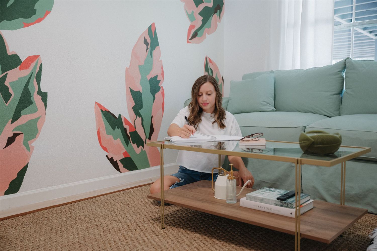 A living room features tropical wall art, mint green sofa, and someone working at a coffee table.