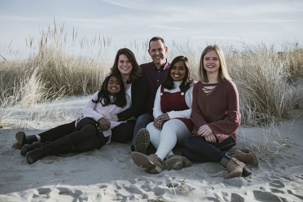 a family of five, a mom, dad, and three daughters on the beach with beach grass in the background