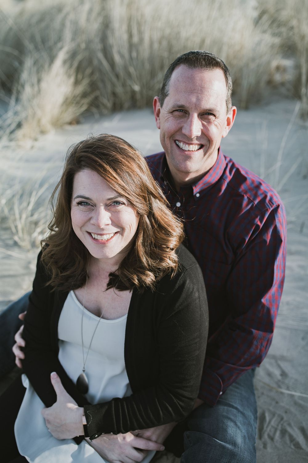 a family photo shoot image of mom and dad, husband & wife, sitting on the beach with cream beach grass in the background